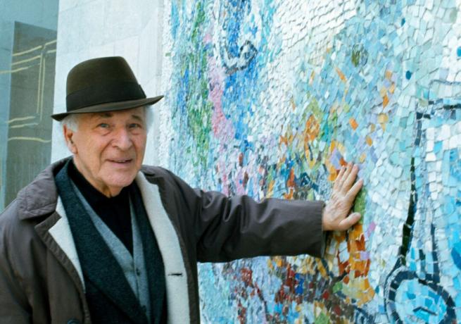 Ο καλλιτέχνης Marc Chagall που φοράει καπέλο πιέζει το χέρι του σε έναν τοίχο με μπλε μωσαϊκά σχέδια.